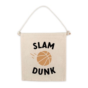 Peut inclure: Une bannière en toile beige avec le texte "SLAM DUNK" en lettres noires. Un ballon de basket brun avec des lignes noires est au centre de la bannière.