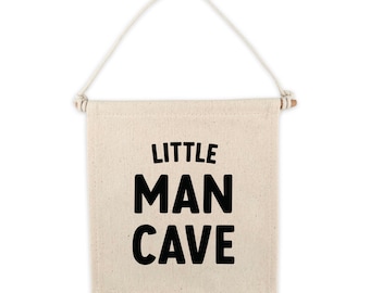 May include: A beige canvas banner with a wooden dowel at the top and a rope hanger. The banner has black text that reads "LITTLE MAN CAVE".