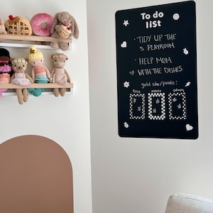 May include: A black to-do list board with white text and decorative elements. The text reads "To do list" followed by "Tidy up the playroom" and "Help mom with the dishes." Below are three sections for gold stars/points, with names listed below each section.