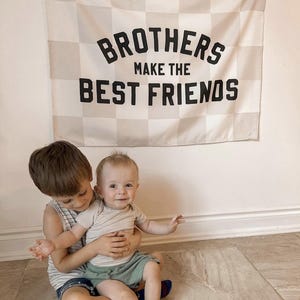 May include: Two brothers, one sitting on the other's lap, in front of a chequered banner that reads "Brothers Make the Best Friends".