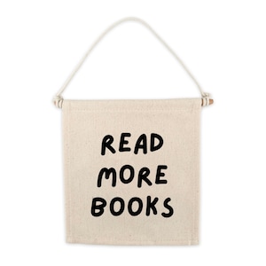 Peut inclure: Une bannière en toile blanche avec un bâtonnet en bois en haut et une attache en corde. La bannière porte un texte noir qui dit "LIRE PLUS DE LIVRES".