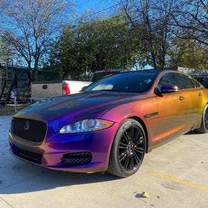 May include: A Jaguar sedan featuring a color-shifting iridescent paint finish that transitions from deep purple at the front to a warm golden orange along the side panels, equipped with black multi-spoke alloy wheels and a black mesh front grille.
