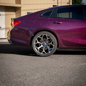 May include: A close-up of the rear side of a dark purple car with a sparkling finish. The car has a silver alloy wheel and a black tire. The car is parked on a gray asphalt surface.