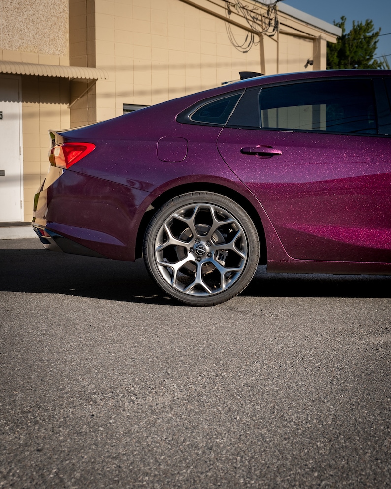 May include: A close-up of the rear side of a dark purple car with a sparkling finish. The car has a silver alloy wheel and a black tire. The car is parked on a gray asphalt surface.