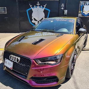 May include: A custom-painted Audi car with a color-shifting finish, transitioning from gold to purple. The car features a black grille, headlights, and a license plate. The car is parked on a paved surface.