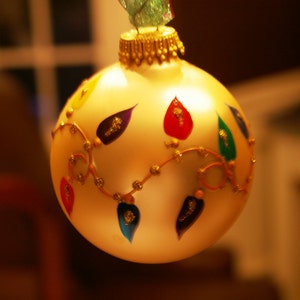 以下が含まれることがあります： 金色の縁取りとカラフルなペイントされたライトが付いた白いクリスマスオーナメント。オーナメントは、金色と白の渦巻き模様で飾られています。