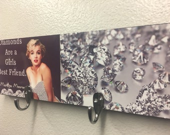 May include: A wooden wall-mounted coat rack with two hooks. The rack features a printed image of diamonds and a quote that reads "Diamonds Are a Girls Best Friend. Marilyn Monroe."