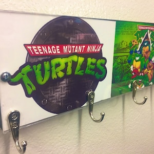 May include: A wooden key holder with a Teenage Mutant Ninja Turtles theme. The holder features a sewer grate with the Teenage Mutant Ninja Turtles logo and a scene from the cartoon. The holder has four metal hooks.