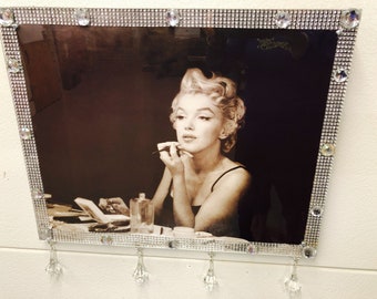 May include: A framed black and white photograph of a woman applying lipstick. The frame is decorated with a silver border and rhinestones.