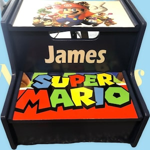 May include: A blue wooden step stool with a personalized name "James" and a Super Mario Bros. themed design. The top step features a colorful image of Mario and other characters from the game. The bottom step features the words "Super Mario" in a colorful, bold font.