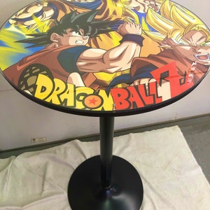 May include: A black round table with a Dragon Ball Z themed tabletop featuring Goku and Vegeta in a battle pose. The table has a black metal base with a round foot.