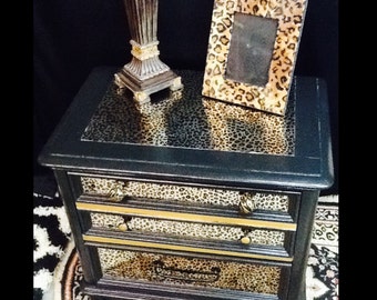 May include: Black bedside table with two drawers featuring a leopard print design. The bedside table has gold trim and gold handles. A small leopard print picture frame sits on top of the bedside table.