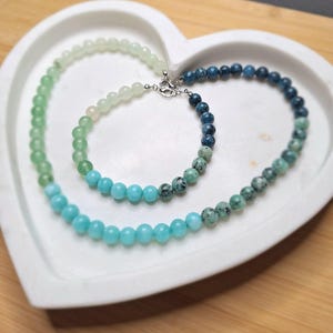 May include: A necklace and bracelet set featuring beaded strands in shades of green and blue. The necklace and bracelet are displayed on a white heart-shaped tray. The beads transition from light to dark colors.