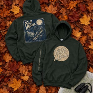 May include: A dark green hooded sweatshirt featuring a Full Moon Hiking Club graphic on the back, a Full Moon circular logo on the front, and the text Hike Wild, Roam Free, Be Legendary printed vertically along the right sleeve.