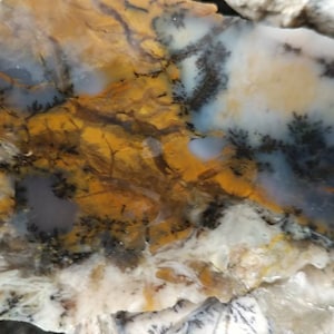 Puede incluir: Una losa de piedra natural con un patrón único. La piedra presenta una mezcla de colores, incluyendo amarillo, blanco, gris y negro. El patrón se asemeja a un paisaje con formaciones arbóreas. Una marca blanca es visible.