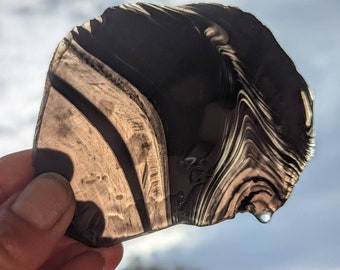 May include: A dark gray, translucent, irregularly shaped piece of glass with a swirling, layered pattern. The glass has a water droplet hanging from the bottom edge.