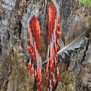 Puede incluir: Un par de aretes largos y abalorios con un esquema de color rojo, naranja y blanco. Los aretes cuelgan de un tronco de árbol.