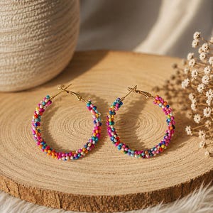 May include: A pair of hoop earrings featuring a circular gold-tone metal frame densely covered in tiny, multi-colored seed beads. The beads display a vibrant mix of pink, blue, yellow, orange, and white colors, creating a textured, colorful surface.