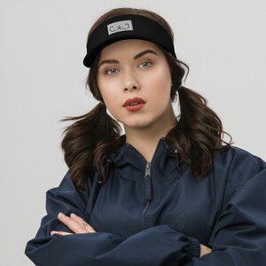 May include: A black visor with a white patch that says "C&C" on it. The visor is being worn by a person with long brown hair pulled back in pigtails. The person is wearing a dark blue jacket.