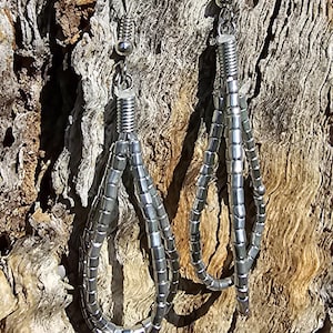 Puede incluir: Un par de pendientes de plata con diseño de cuentas. Los pendientes están hechos de pequeñas cuentas redondas que están enhebradas juntas en un patrón largo y fluido. Los pendientes cuelgan de un gancho de plata.