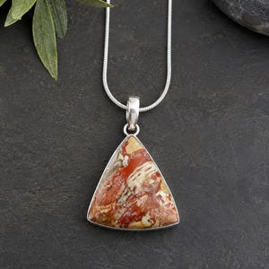 May include: A triangular pendant necklace featuring a polished, multi-colored stone with swirling patterns of red, cream, and brown. The stone is set in a silver-toned metal frame and hangs from a matching silver-toned snake chain necklace.