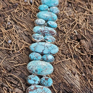 May include: A turquoise and silver bracelet with a row of oval-shaped turquoise stones. The stones are a light blue color with brown and black markings. The bracelet is made of sterling silver and has a clasp.