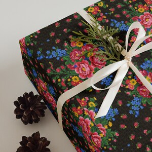 Può includere: Una scatola regalo nera avvolta in carta con motivo floreale con un nastro di raso bianco legato in un fiocco. Un piccolo rametto di verde è infilato sotto il nastro.