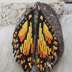 Puede incluir: Un par de pendientes largos de cuentas con un esquema de color negro, naranja y amarillo. Los pendientes están hechos de pequeñas cuentas redondas y tienen un diseño de flecos.
