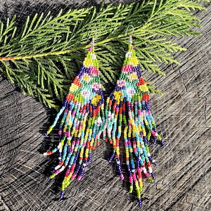 Puede incluir: Pendientes de cuentas coloridas con un diseño triangular y flecos largos. Los pendientes presentan una vibrante mezcla de colores, incluyendo azul, verde, amarillo, rojo y rosa. Los pendientes se exhiben sobre una superficie de madera con una rama verde.