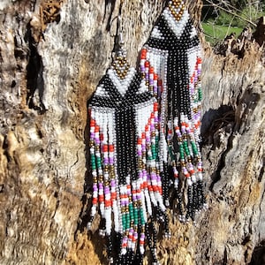 Puede incluir: Un par de pendientes largos de cuentas con un patrón geométrico en negro, blanco, dorado, rojo, verde y morado. Los pendientes están colgando de un tronco de árbol.