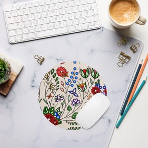 Puede incluir: Alfombrilla de ratón redonda beige con un diseño floral colorido con flores y hojas rojas, azules y verdes. Un ratón de ordenador blanco descansa sobre la alfombrilla, con un teclado y una taza de café en el fondo.