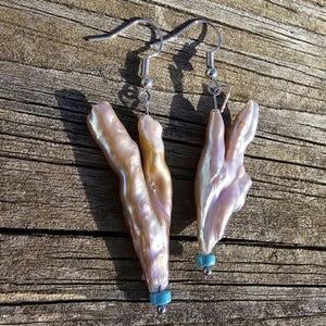 May include: A pair of earrings featuring iridescent baroque pearls with a turquoise accent bead on each earring. The pearls are a light pink and cream color with a pearlescent sheen. The earrings are attached to silver hooks.