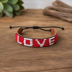 Pode incluir: Uma pulseira de contas com base de couro e contas de sementes vermelhas e brancas que formam a palavra LOVE. A pulseira inclui um fecho de cordão preto com nós deslizantes ajustáveis, destacando o contraste entre as contas e o couro.