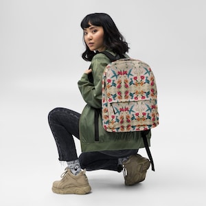 May include: A beige backpack with a floral pattern featuring hummingbirds and flowers. The backpack has black straps and a black zipper.