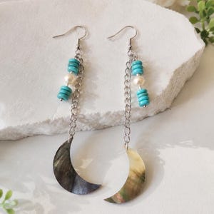 May include: A pair of dangle earrings featuring crescent moon pendants. The earrings have silver-tone chains, turquoise beads, and white pearl beads. The moon pendants have a pearlescent finish, with shades of gray, brown, and gold.