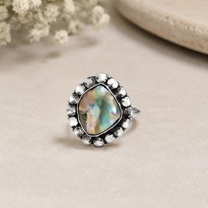 May include: A silver ring featuring a large, iridescent, square-shaped stone. The stone is surrounded by a border of small, round silver accents. The ring's band is a simple, thin silver design. The ring is set against a neutral background.