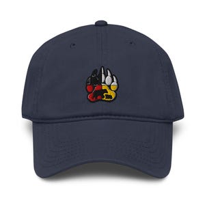 May include: Navy blue baseball cap with a colorful embroidered bear paw design. The paw print features black, red, yellow, and white accents, with a bear silhouette in the center. A classic, adjustable hat.