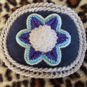 May include: A handcrafted, beaded flower pin. The pin features a navy blue base with a beaded flower in shades of blue and turquoise, and a white beaded center. The edges are trimmed with a delicate, clear beaded border.