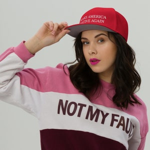 May include: A red and gray baseball cap with the text "Make America Native Again" in white. The person wearing the cap is also wearing a pink, white, and burgundy striped sweater with the text "Not My Fault" printed on it.