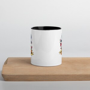 Könnte beinhalten: Weiße Keramiktasse mit schwarzem Innenraum und einem farbenfrohen Design im Tattoo-Stil. Das Design umfasst einen blauen Vogel, rote Blumen und ein gelbes Banner. Die Tasse steht auf einem Holzschneidebrett.