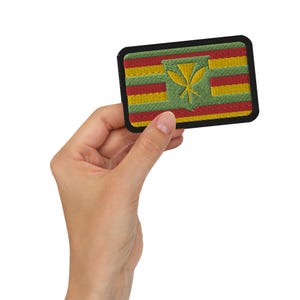 May include: Embroidered patch featuring the Hawaiian flag design. The patch has a black border, with a green background and red, yellow, and green stripes. A yellow emblem is centered on the patch.