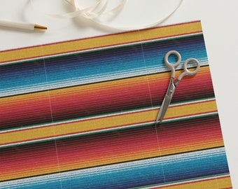 Puede incluir: Una tela de rayas coloridas con un patrón tradicional de sarape mexicano en rojo, amarillo, azul y blanco. Un par de tijeras plateadas está sobre la tela.