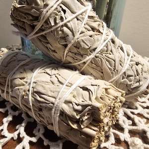 Puede incluir: Dos manojos de varitas de salvia blanca atadas con cuerda blanca. La salvia está seca y atada en manojos, y las varitas están listas para ser quemadas para la limpieza.