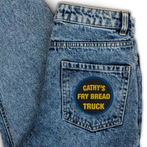 Può includere: Un paio di jeans blu denim con una toppa ricamata nera e gialla sulla tasca posteriore che dice "Cathy's Fry Bread Truck".