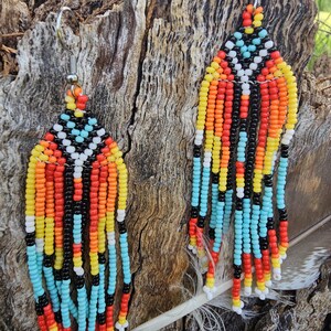 Puede incluir: Un par de aretes largos y abalorios con un patrón geométrico en tonos de rojo, naranja, amarillo, azul, negro y blanco. Los aretes cuelgan de ganchos plateados.