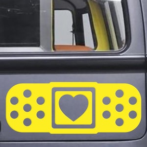 May include: A yellow bandage sticker with a gray heart design on the back of a gray van. The sticker has a rectangular shape with a heart in the center and dotted edges. The van has a roof rack.