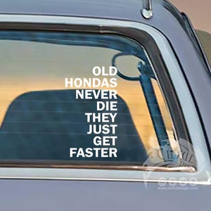 May include: A close-up of the rear window of a grey vintage Honda car. Two white decals read "OLD HONDAS NEVER DIE THEY JUST GET FASTER". The car has a red tail light and a chrome bumper.