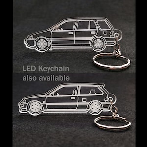 May include: A white outline of a car keychain with a silver metal ring. The keychain is made of clear acrylic and features a detailed outline of a car. The text "LED Keychain also available" is printed below the keychain.