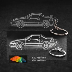 Puede incluir: Un llavero de acrílico negro con un contorno blanco de un coche Honda CRX. El llavero tiene un anillo y una cadena de metal plateado. El texto "LED keychain also available" está impreso debajo del coche.