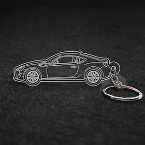 May include: A white outline of a car keychain with a silver key ring. The car is a coupe with a sleek design. The keychain is on a black background.